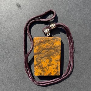 May include: A rectangular pendant with a mottled orange and brown pattern, suspended from a dark purple cord necklace. The cord is attached to the pendant with silver-colored metal accents.