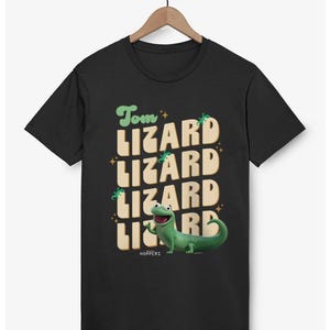 May include: Black t-shirt with the text "Tom Lizard Lizard Lizard Lizard" in a retro font. A green lizard illustration is on the bottom right. The shirt is hanging on a wooden hanger.