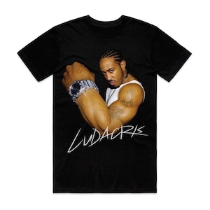 May include: Black t-shirt featuring a portrait of a man flexing his arm, wearing a silver watch and a white tank top. The word "Ludacris" is written in white script across the bottom of the image.