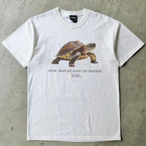 Puede incluir: Una camiseta blanca de manga corta con un gráfico de una tortuga marrón y beige en el centro. Debajo de la tortuga, el texto dice "slow motion over no motion" seguido del logotipo de National Geographic en letras negras y amarillas.