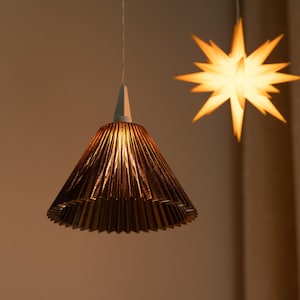 Könnte beinhalten: Ein kupferfarbener, plissierter Lampenschirm hängt an einer weißen Halterung. Der Lampenschirm hat eine konische Form und ist von innen beleuchtet. Im Hintergrund befindet sich eine sternförmige Leuchte mit warmem, gelbem Schein. Die Leuchten sind an der Decke aufgehängt.