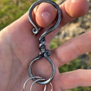 Könnte beinhalten: Handgeschmiedeter Metall-Schlüsselanhänger mit einem einzigartigen, wirbelnden Design. Der Schlüsselanhänger hat oben eine Hakenform, einen verdrehten Mittelteil und unten eine Schlaufe mit zwei silbernen Schlüsselringen. Das Metall hat eine dunkle, strukturierte Oberfläche.