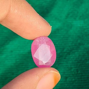 May include: A faceted, oval-shaped, pink gemstone is held between two fingers against a green background. The gem has a polished surface with visible internal facets, reflecting light. The color is a vibrant, saturated pink.