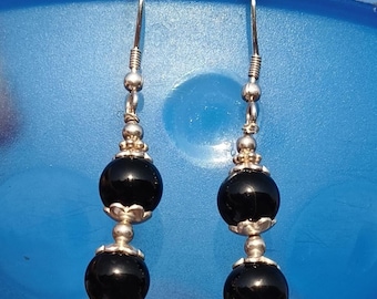 May include: A pair of dangle earrings featuring two polished black beads and silver-colored metal accents. The earrings have a hook closure and are displayed against a blue background. The earrings are a classic jewelry accessory.
