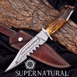 May include: A stainless steel knife with a serrated edge and a deer antler handle, resting in a brown leather sheath. The blade has etched text and a pentagram symbol below the text "SUPERNATURAL".