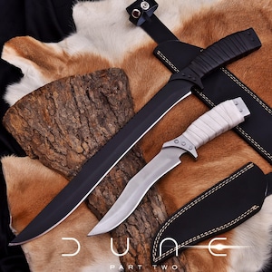 May include: Two knives with sheaths are displayed on a textured surface. One is a large black blade with a leather-wrapped handle. The other is a smaller knife with a silver blade and a white wrapped handle. The text "DUNE PART TWO" is at the bottom.