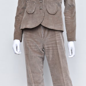 May include: A brown corduroy suit comprising a jacket and trousers. The jacket features two rounded pockets and three buttons. The trousers are straight-legged. The suit is displayed on a white mannequin.