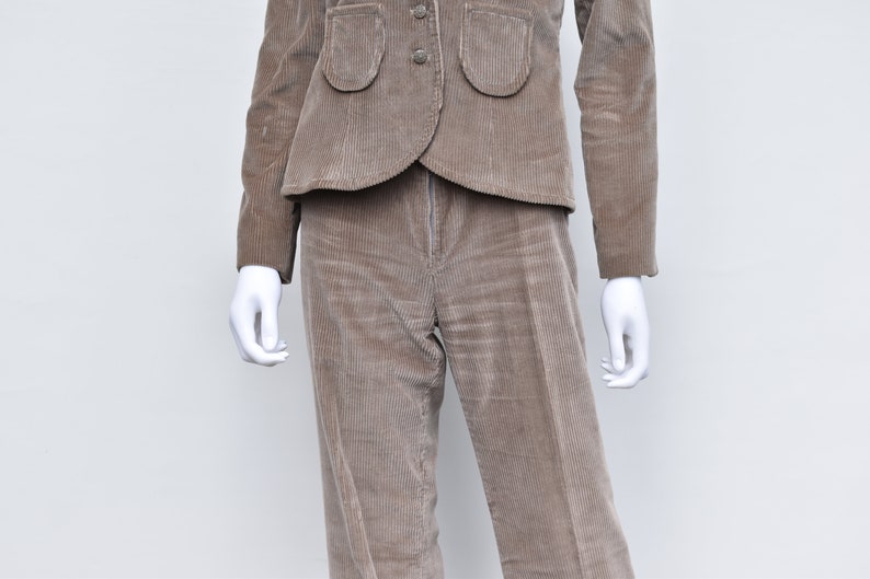 May include: A brown corduroy suit comprising a jacket and trousers. The jacket features two rounded pockets and three buttons. The trousers are straight-legged. The suit is displayed on a white mannequin.