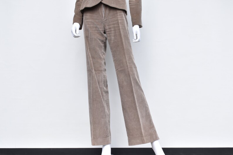 May include: A taupe corduroy suit on a mannequin. The suit comprises a blazer and wide-leg trousers. The fabric has a vertical ribbed texture. The mannequin is set against a plain white background.