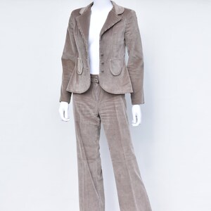 May include: A taupe corduroy suit on a mannequin. The jacket has a notched lapel, three buttons, and two pockets. The trousers are flared and have a belt. The suit is a neutral colour, suitable for various occasions. The suit is made of corduroy.