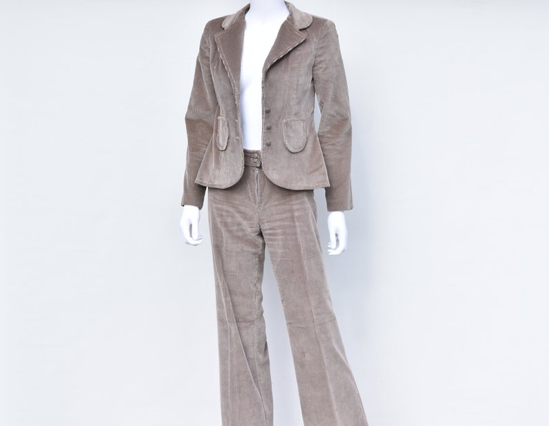 May include: A taupe corduroy suit on a mannequin. The jacket has a notched lapel, three buttons, and two pockets. The trousers are flared and have a belt. The suit is a neutral colour, suitable for various occasions. The suit is made of corduroy.