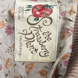 May include: Close-up of a label from "The Strawberry Patch" by Dorothy Schoelen, California. The label is rectangular with a cream background and embroidered text and a strawberry illustration. The background fabric has a floral pattern.