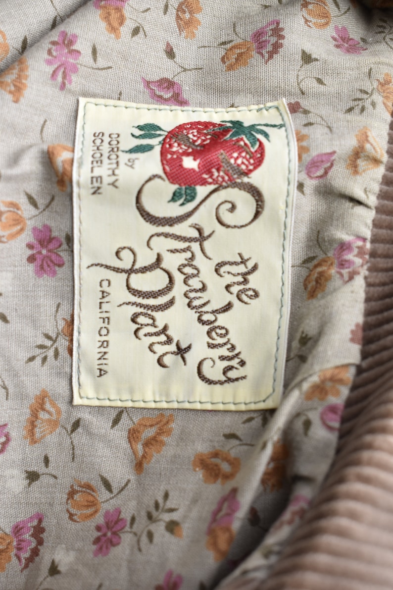May include: Close-up of a label from "The Strawberry Patch" by Dorothy Schoelen, California. The label is rectangular with a cream background and embroidered text and a strawberry illustration. The background fabric has a floral pattern.
