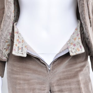 May include: A tan corduroy suit, comprising a jacket and trousers, displayed on a mannequin. The jacket features a floral patterned lining. The trousers have a zip closure. The suit is in a neutral colour.