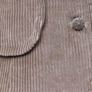 May include: Close-up of a corduroy jacket in a muted taupe colour. The jacket features a rounded pocket detail and a decorative silver button. The fabric has a ribbed texture.