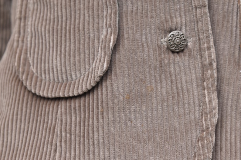 May include: Close-up of a corduroy jacket in a muted taupe colour. The jacket features a rounded pocket detail and a decorative silver button. The fabric has a ribbed texture.