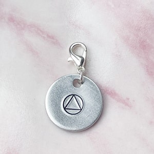 May include: Silver charm with a circle design featuring a triangle inside. The charm has a lobster clasp.