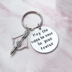 Peut inclure: Porte-clés argenté avec un charme en forme d'arc et de flèche et une étiquette ronde qui dit "Que les chances soient toujours en votre faveur".