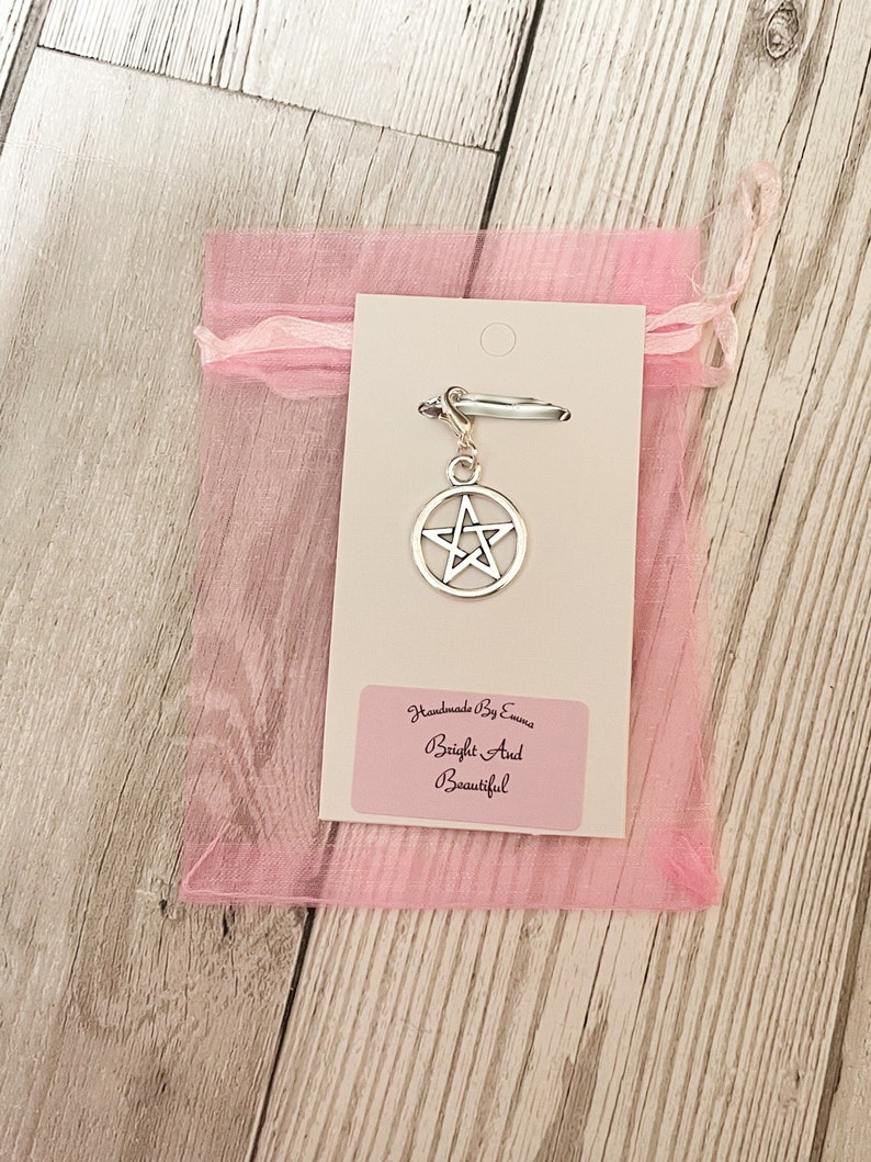 May include: A silver-colored pentagram charm hangs from a silver clasp, attached to a white card. The card is inside a sheer pink drawstring bag. The text on the card reads "Handmade by Emma, Bright and Beautiful."