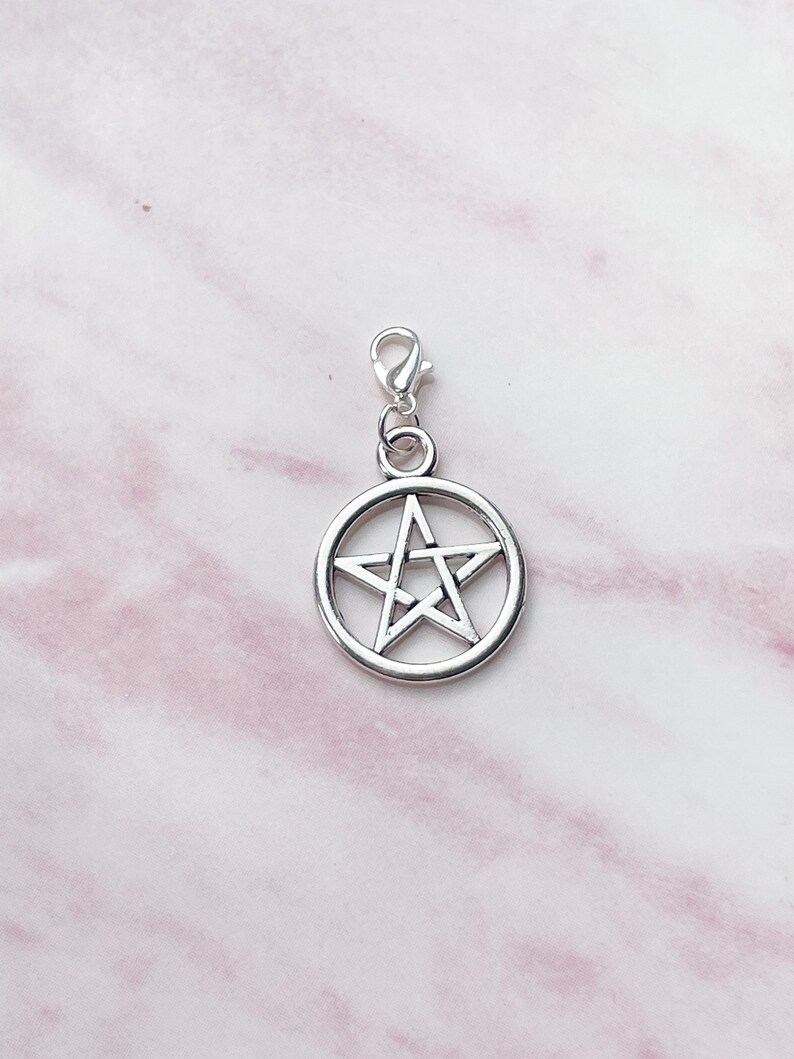 May include: A silver-colored pentagram charm with a lobster clasp. The five-pointed star is set within a circular frame. The charm is shown against a pink and white marble background.