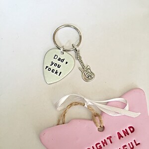 May include: A silver guitar pick keychain with the text "Dad, you rock!" and a small silver guitar charm. A pink ceramic butterfly ornament with the text "Bright and Beautiful" is also shown.