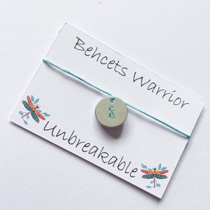May include: A white card with the text "Behcet's Warrior Unbreakable" and a teal blue string with a silver charm with a teal blue ribbon on it. There are two dragonfly illustrations on the card.