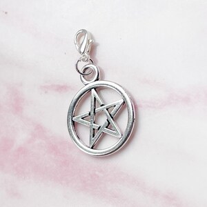 May include: Silver-colored pentagram charm with a lobster clasp. The five-pointed star is encircled by a ring. The charm is designed for jewelry making or as a pendant. The background is a light pink and white fabric.