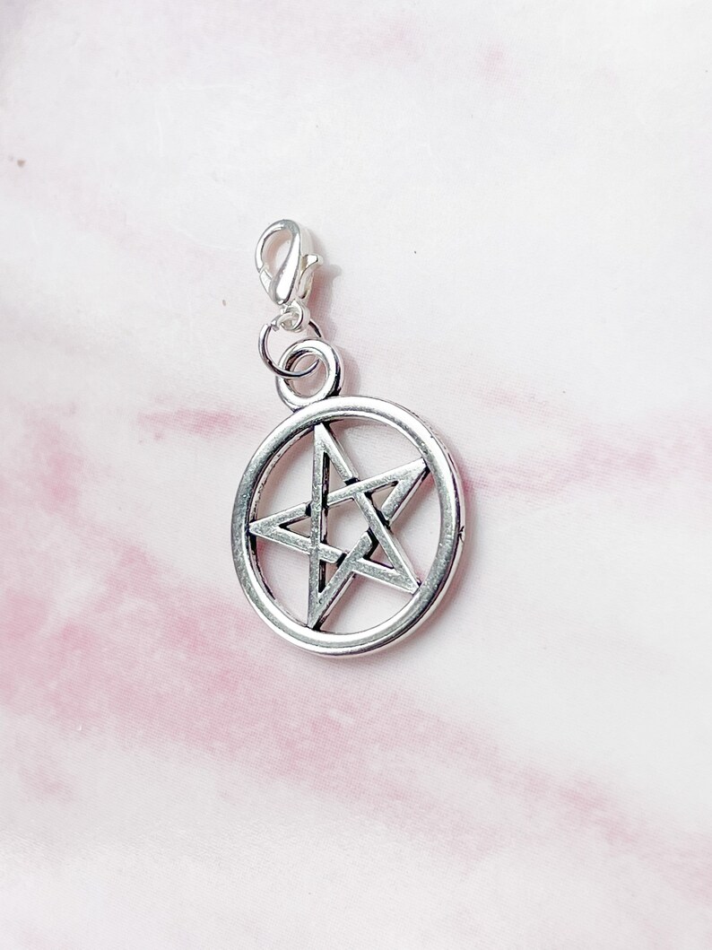 May include: Silver-colored pentagram charm with a lobster clasp. The five-pointed star is encircled by a ring. The charm is designed for jewelry making or as a pendant. The background is a light pink and white fabric.