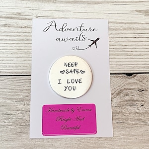 May include: A white card with the words "Adventure awaits" and a small airplane illustration. A silver-colored circular charm is stamped with "KEEP SAFE I LOVE YOU." A pink label reads "Handmade by Emma Bright And Beautiful."