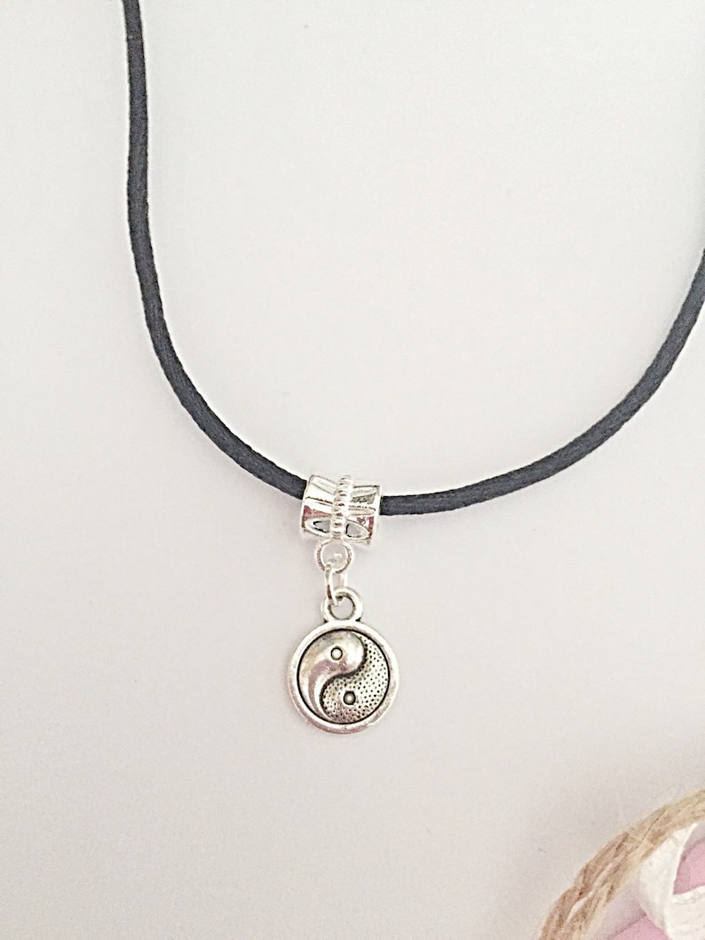 Może przedstawiać: Czarny sznurek z naszyjnikiem z srebrnym symbolem yin yang. Zawieszka jest przymocowana do sznurka srebrną koralikiem.