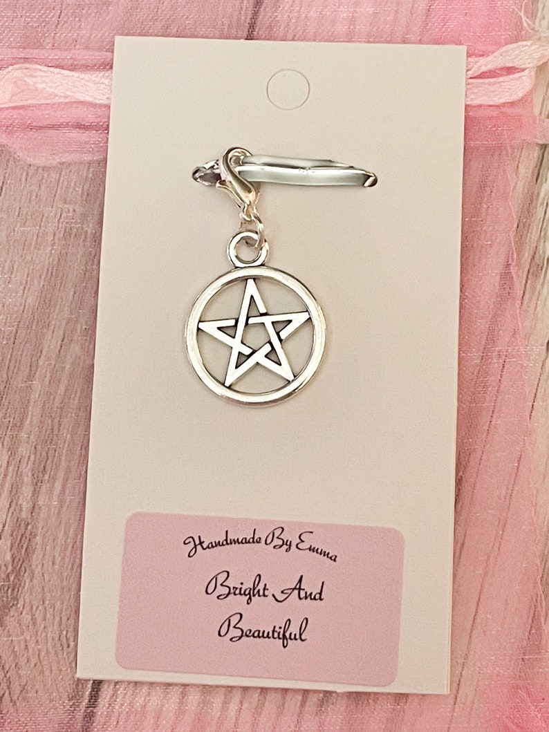 May include: A silver-colored charm with a pentagram design, attached to a clasp. The charm is on a white card with a pink label that reads "Handmade By Emma Bright And Beautiful". The background is a soft pink.