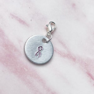 May include: Silver charm with a pink ribbon design, a symbol of breast cancer awareness. The charm has a lobster clasp for attaching to a bracelet or necklace.