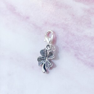 May include: Silver charm with a four-leaf clover design. The charm has a lobster clasp for attaching to a bracelet or necklace.