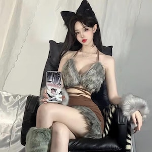 Puede incluir: Un conjunto marrón de top halter y falda con ribete de piel sintética. El conjunto incluye puños y calentadores de piernas a juego de piel sintética. Se llevan orejas de gato negras en la cabeza. Un teléfono con un gráfico de manga se sostiene en la mano.