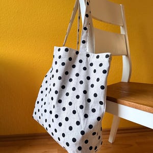 Könnte beinhalten: Eine graue Tragetasche mit schwarzen Punkten hängt an einem weißen Stuhl. Die Tasche hat lange Träger und ist aus einem hellen Stoff gefertigt. Der Hintergrund ist eine gelbe Wand und ein Holzboden.