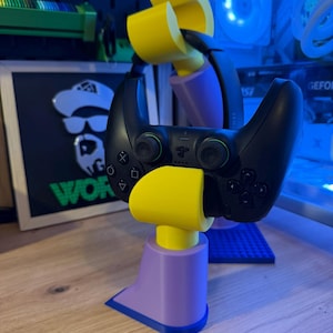 Può includere: Un controller di gioco nero è tenuto da un supporto stampato in 3D. Il supporto è composto da componenti gialli e viola. Il controller ha pulsanti e joystick. Il supporto è su una superficie di legno.