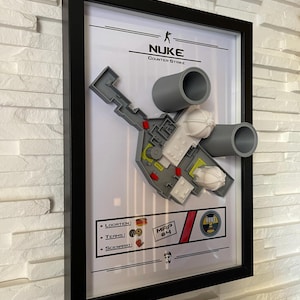 Puede incluir: Obra de arte enmarcada que presenta un modelo 3D del mapa "Nuke" de Counter-Strike. El mapa es gris, blanco y amarillo, con dos estructuras cilíndricas grises. La obra, enmarcada en negro, incluye el logotipo del juego y detalles del mapa.