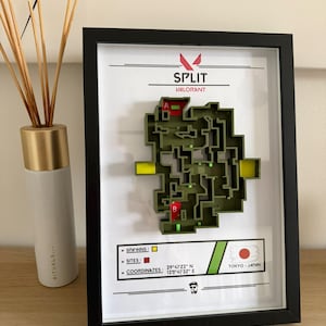 Peut inclure: Une carte 3D encadrée de la carte "Split" du jeu Valorant. La carte est verte avec des marqueurs jaunes et rouges, sur fond blanc. Le cadre est noir, et le texte "Split" et "Valorant" est visible.
