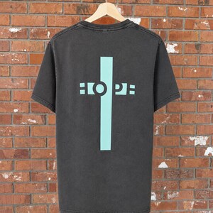 May include: A charcoal grey short-sleeve t-shirt featuring a large, light blue vertical cross graphic on the back. The word HOPE is printed in light blue capital letters across the horizontal bar of the cross, with the O integrated into the design.