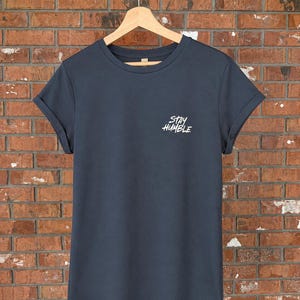 May include: A dark charcoal grey short-sleeve t-shirt with a crew neckline and rolled sleeves. The front of the shirt features the white text "STAY HUMBLE" printed in a stylised font on the upper left chest area, hanging on a wooden hanger.