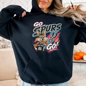May include: Black hoodie with a cartoon coyote wearing a basketball jersey with the number 21. The hoodie displays the words "GO SPURS GO!" in a colourful design. The hood is up.