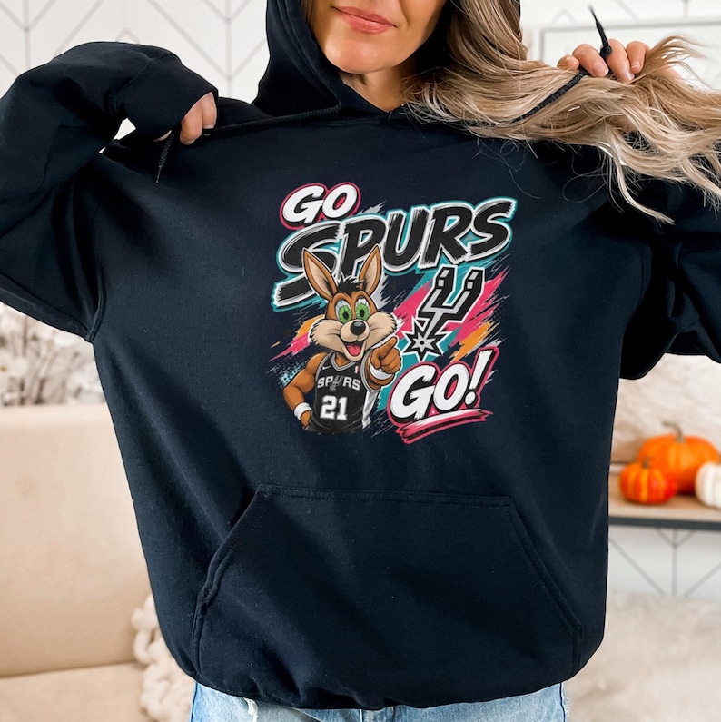 May include: Black hoodie with a cartoon coyote wearing a basketball jersey with the number 21. The hoodie displays the words "GO SPURS GO!" in a colourful design. The hood is up.