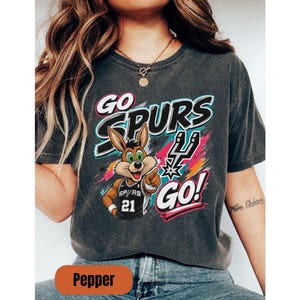 May include: Dark grey t-shirt with the text "GO SPURS GO!" and a cartoon rabbit in a basketball jersey. The jersey number is 21. The design incorporates the Spurs logo and vibrant colour accents.