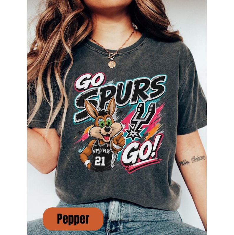 May include: Dark grey t-shirt with the text "GO SPURS GO!" and a cartoon rabbit in a basketball jersey. The jersey number is 21. The design incorporates the Spurs logo and vibrant colour accents.