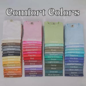 May include: A display of Comfort Colors t-shirts in a variety of shades, such as white, pink, green, and blue. Each stack shows a folded t-shirt with a colour name label underneath. The image presents a wide spectrum of colours.