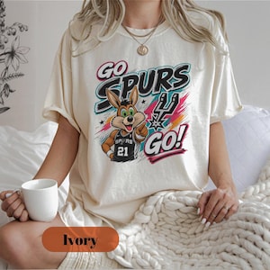 May include: Ivory-coloured t-shirt with a cartoon rabbit wearing a basketball jersey. The text "GO SPURS GO!" is in a colourful design. The rabbit is pointing. The shirt has a relaxed fit. The word "Ivory" is at the bottom.