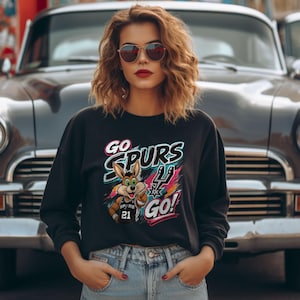 May include: Black crew neck sweatshirt featuring the text "GO SPURS GO!" and a cartoon coyote graphic. The coyote wears a Spurs jersey with the number 21. The design has a colourful, dynamic style.