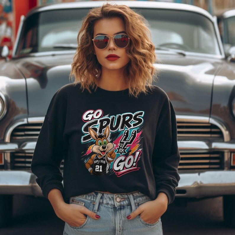 May include: Black crew neck sweatshirt featuring the text "GO SPURS GO!" and a cartoon coyote graphic. The coyote wears a Spurs jersey with the number 21. The design has a colourful, dynamic style.