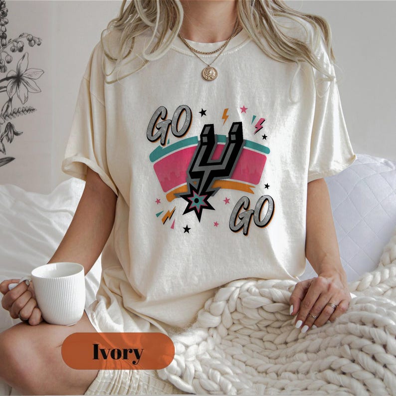 Puede incluir: Camiseta color marfil con un dise&ntilde;o gr&aacute;fico. El dise&ntilde;o incluye las palabras "GO GO" en gris, una pancarta rosa y turquesa y un logotipo de espuela negro y plateado. La camiseta tiene un corte holgado.
