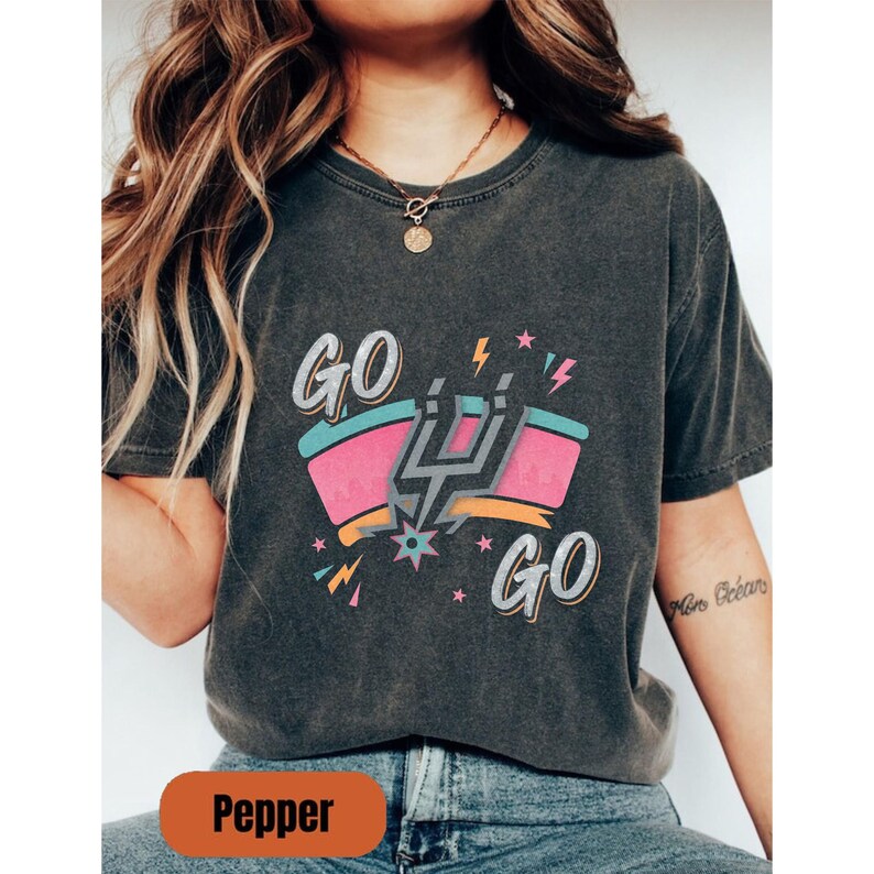 Puede incluir: Camiseta gris oscuro con un dise&ntilde;o gr&aacute;fico colorido. El dise&ntilde;o presenta las palabras "GO GO" con un emblema estilizado en el centro, que incorpora colores rosa, turquesa y naranja. La camiseta tiene un aspecto vintage y desgastado.